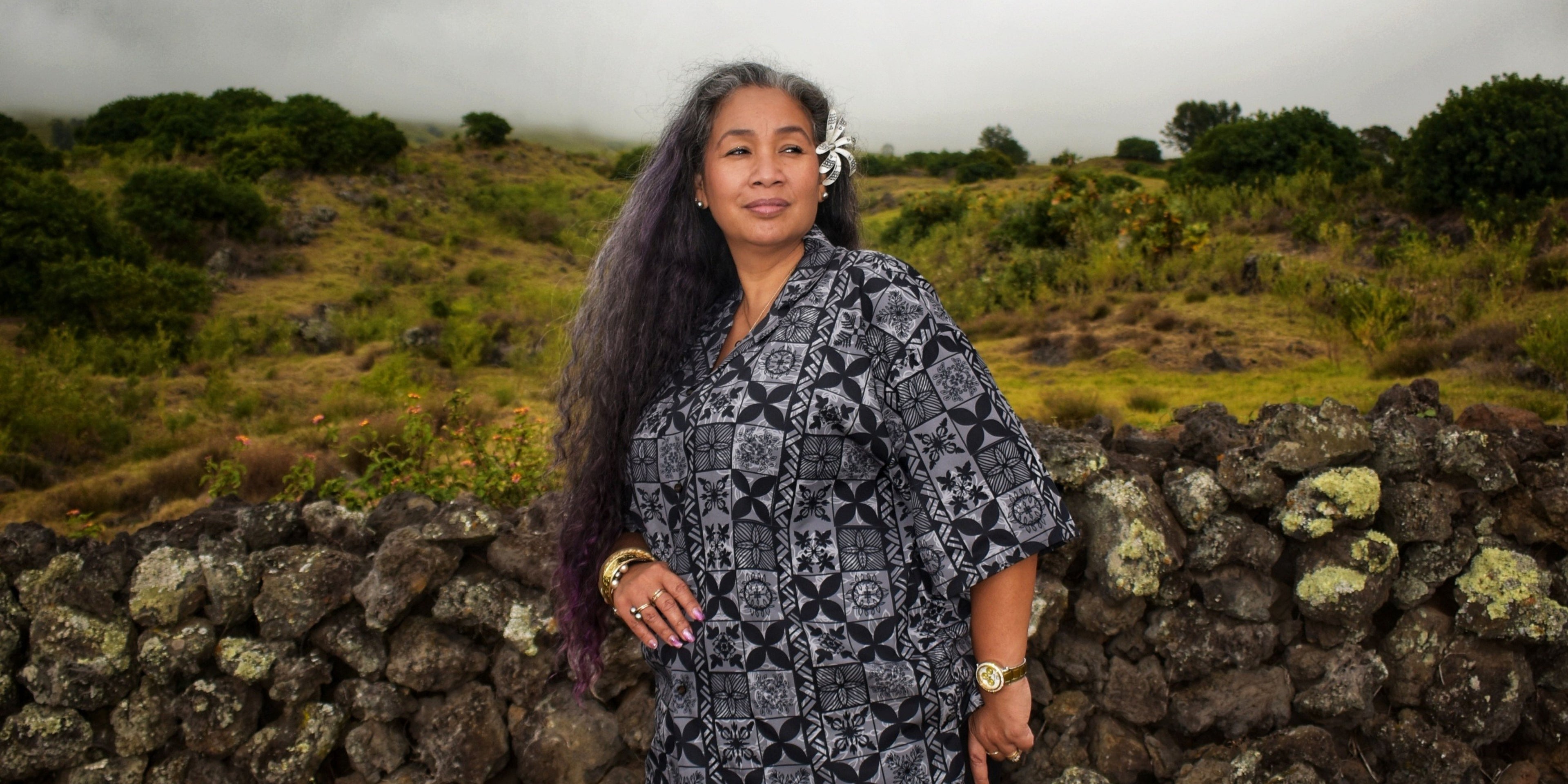 Woman standing in a scenic outdoor setting with a stone wall and greenery.