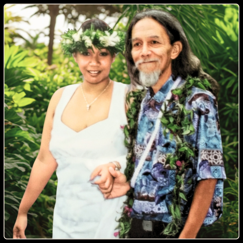 A couple standing together outdoors in front of lush green tropical foliage. The woman wears a white sleeveless dress and a green floral lei, while the man wears a blue patterned short-sleeve shirt and a matching green floral lei.
